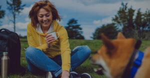 A woman sitting outside with her dog drinking from a metal mug.